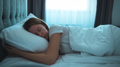 Uma mulher é mostrada dormindo em uma cama. Há uma janela atrás da cama com as cortinas abertas e a luz entrando.