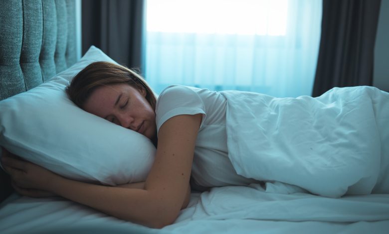 Uma mulher é mostrada dormindo em uma cama. Há uma janela atrás da cama com as cortinas abertas e a luz entrando.