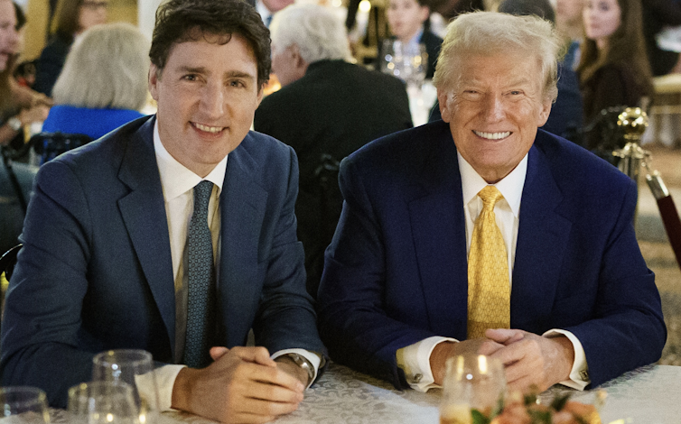 Um homem de cabelos escuros e um homem mais velho com cabelos loiros grisalhos sorriem à mesa de jantar.