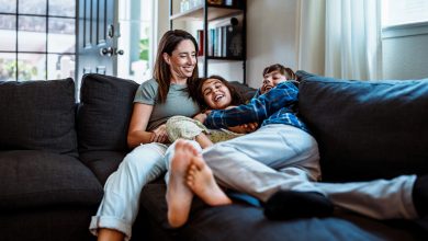 Uma mulher é mostrada deitada no sofá com seus dois filhos. Eles estão todos rindo.