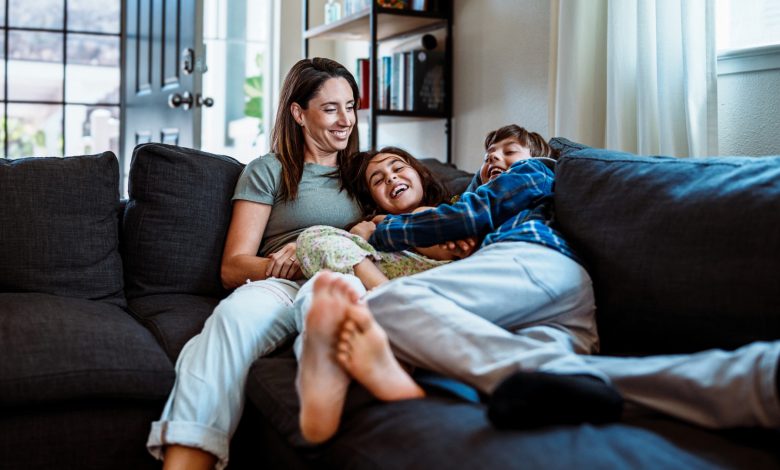 Uma mulher é mostrada deitada no sofá com seus dois filhos. Eles estão todos rindo.