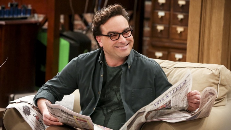 Leonard lendo um jornal em uma poltrona e sorrindo sobre The Big Bang Theory