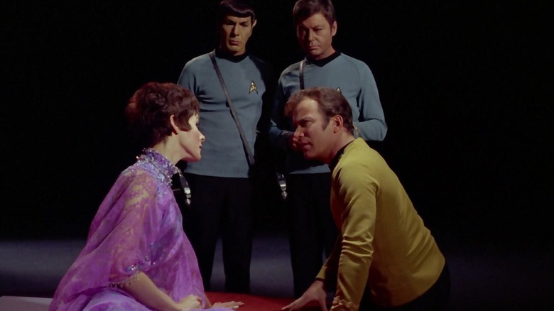 Kirk sentado em uma plataforma com uma mulher misteriosa enquanto Spock e Bones observam em Star Trek