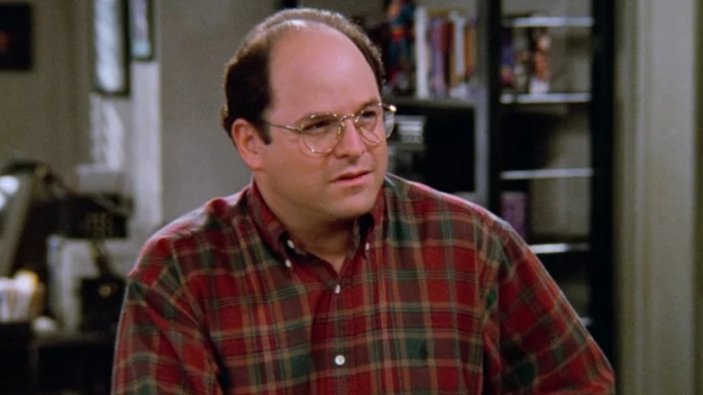 George Costanza, de Jason Alexander, vestindo xadrez, parado no apartamento de Jerry em Seinfeld